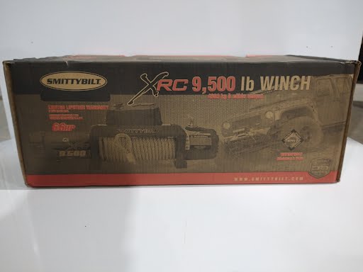 Electric Smittybilt 9500lb winch from PJ Trailers MFG, Inc., featuring a compact design with a steel cable and control switch.